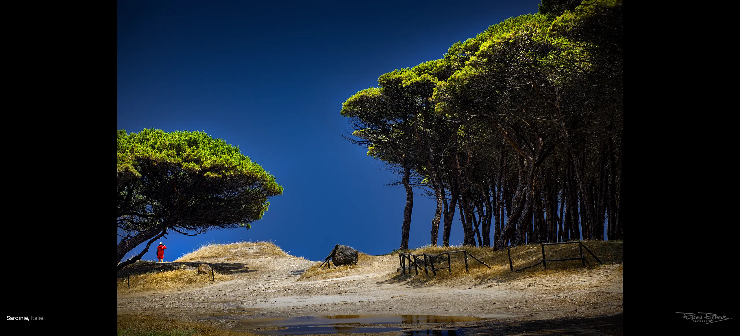 Foto van vrouw in rode jas onder groene bomen tegen blauwe lucht in Sardinië Italië, te zien in rubriek Ruden Riemens Photography Onderweg op website www.rudenriemens.com.