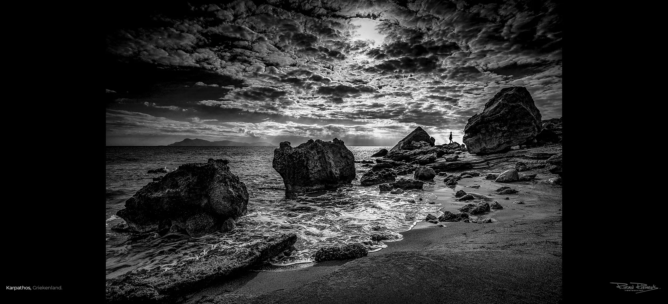 Ruden Riemens fotografeerde aan zee een meisje op de rotsachtige kust van Karpathos Griekenland in zwart-wit onder een prachtige wolkenlucht.