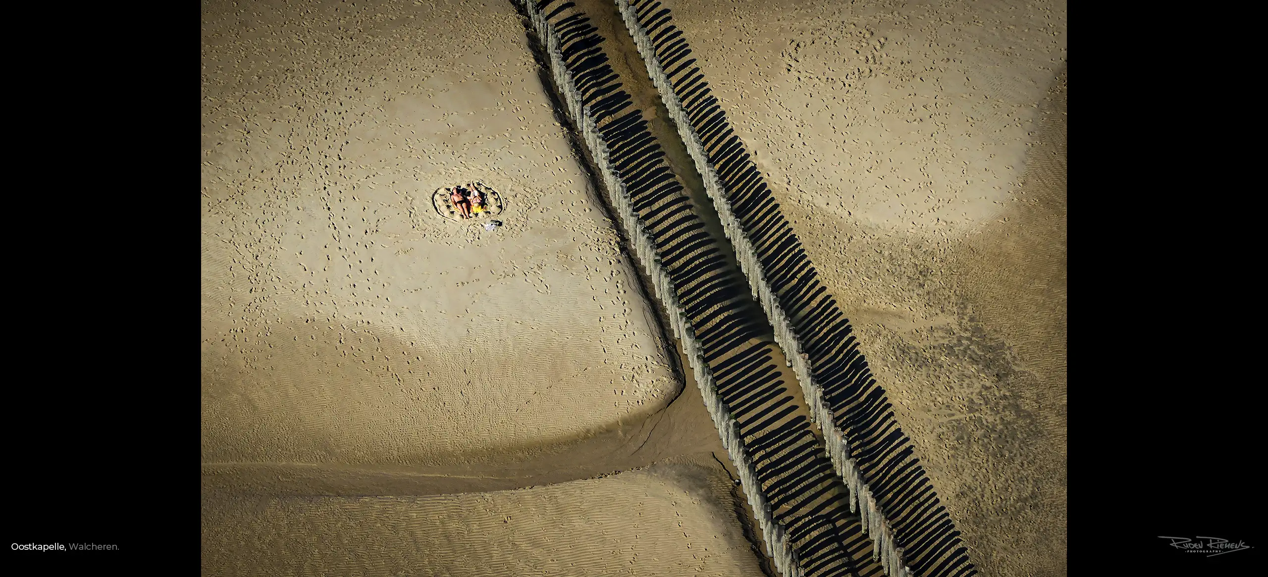 Luchtfoto van een zonnend echtpaar in de zomerzon bij een paalhoofd op het strand van Oostkapelle, gefotografeerd door Ruden Riemens Fotograaf, www.rudenriemens.com.