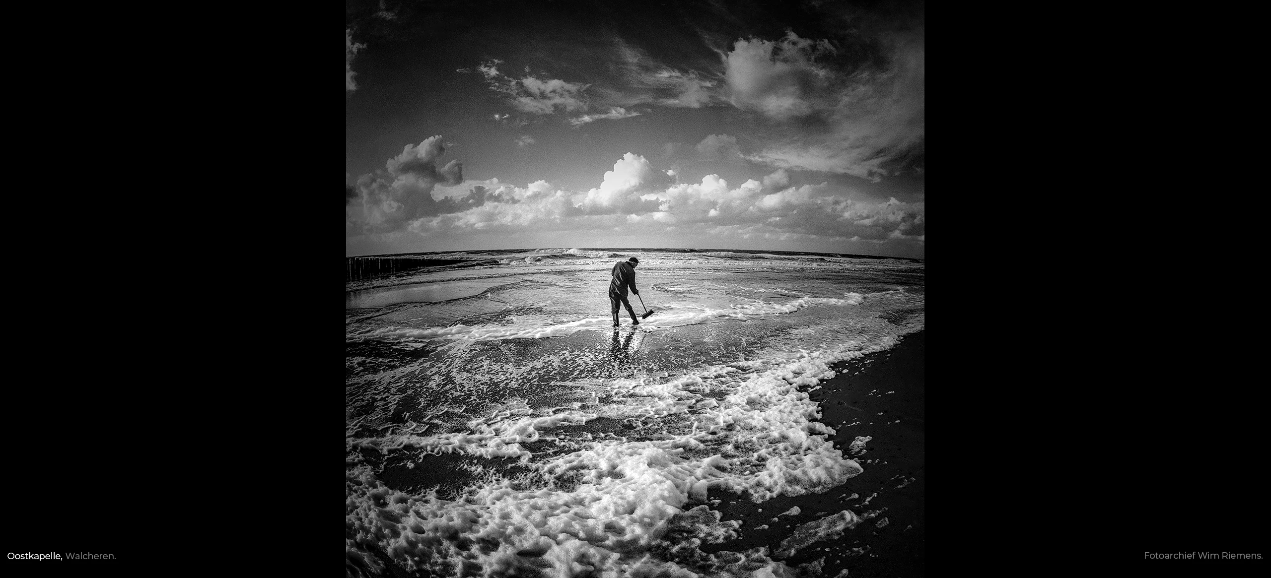 Man met bezem maakt zijn laarzen schoon in schuimende zee onder wolkenlucht te Oostkapelle in Zeeland, foto Wim Riemens.