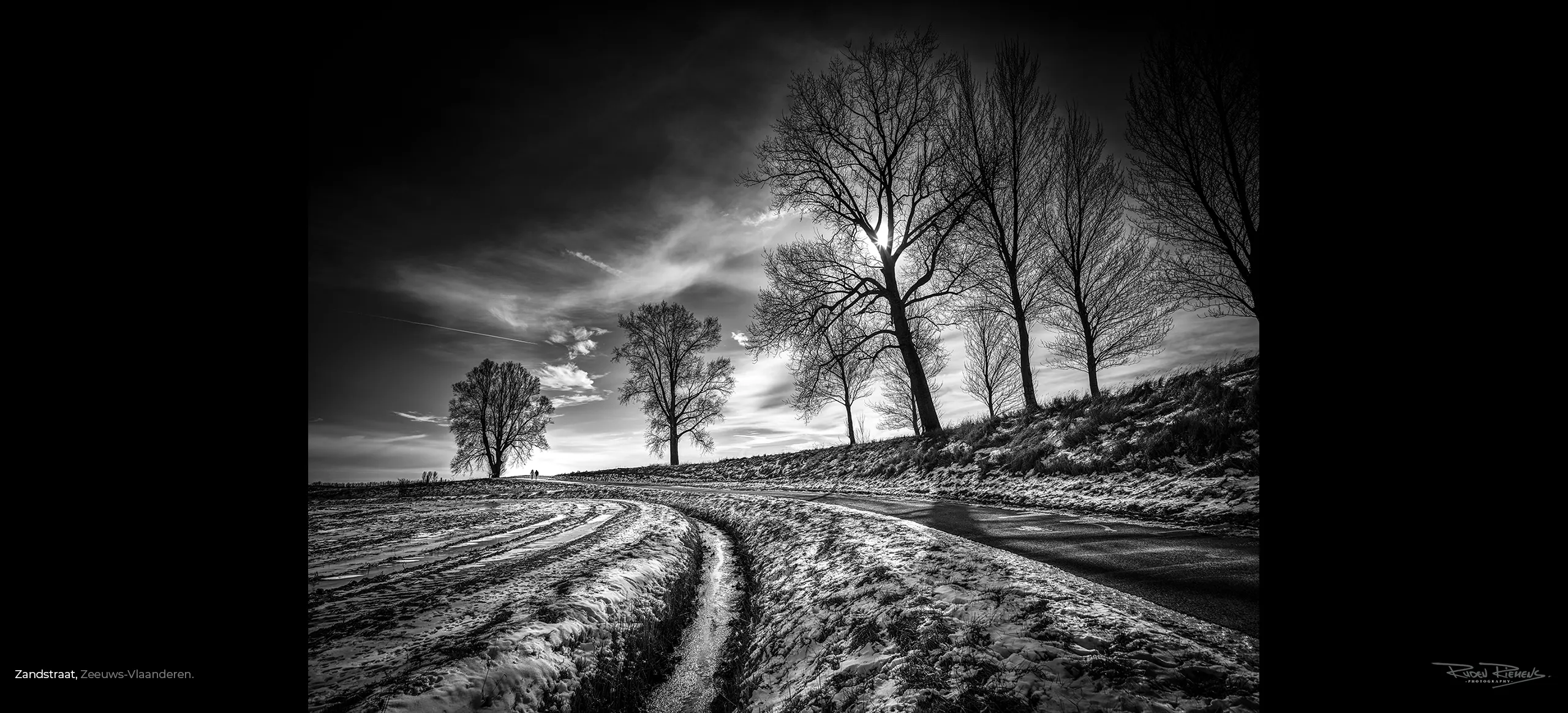 Zwart-wit foto van boomdijk met twee wandelaars in de winter omgeving Zandstraat in Zeeuws-Vlaanderen, gefotografeerd door de Zeelandfotograaf Ruden Riemens.