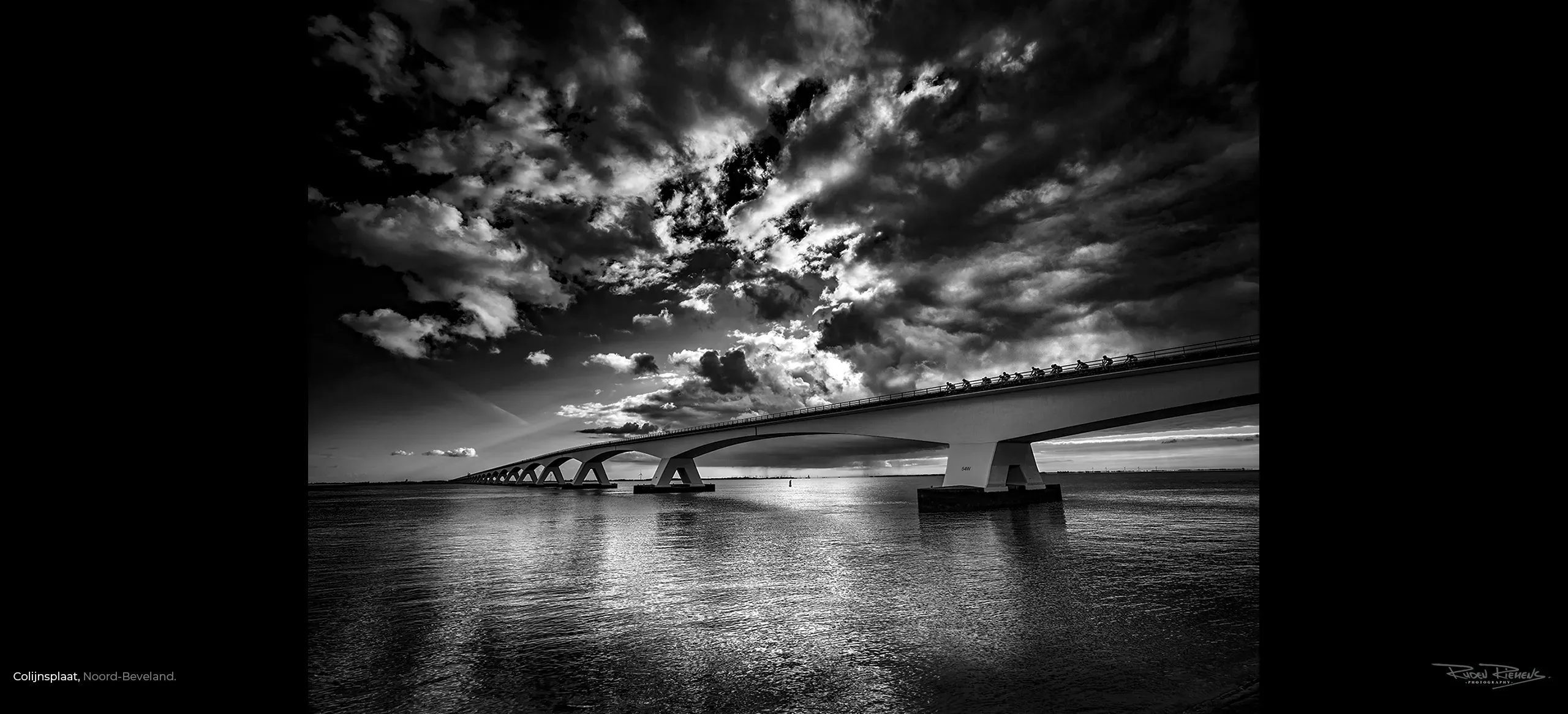 Zwart-wit foto van groepje wielrenners op de Zeelandbrug over de Oosterschelde onder een dreigende wolkenlucht. Ruden Riemens fotografeerde en schreef er iets bij.